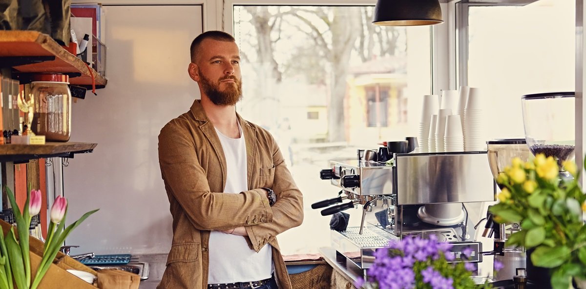 A man in a small cafe with a lot of flowers and coffee machine.