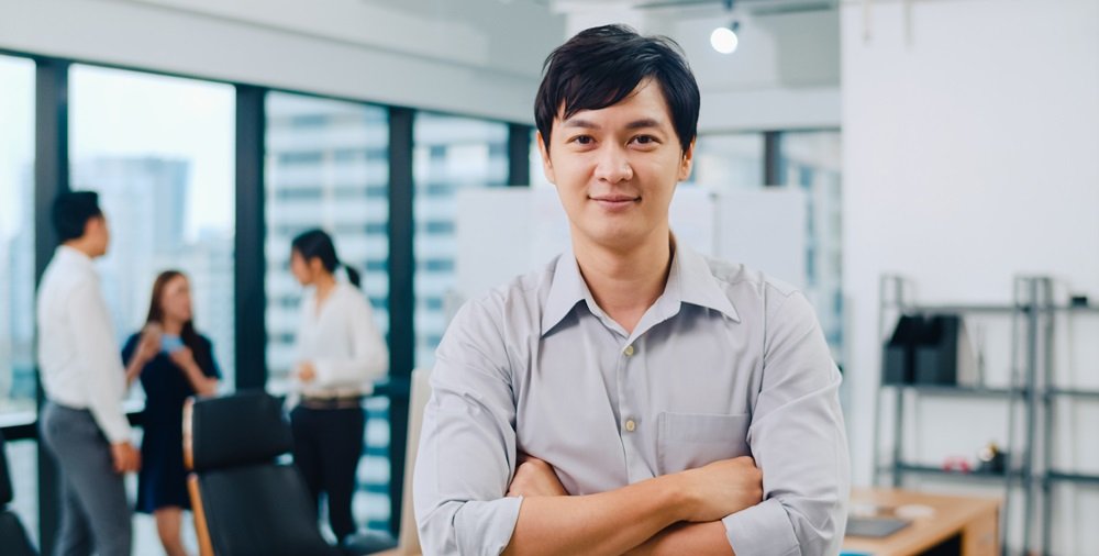 Portrait of successful handsome executive businessman smart casual wear looking at camera and smiling, arms crossed in modern office workplace. Young Asia guy standing in contemporary meeting room.